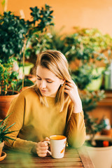 Wall Mural - Romantic portrait of beautiful young woman wearing brown cotton dress, relaxing on the balcony between many green plants, holding cup of tea or coffee