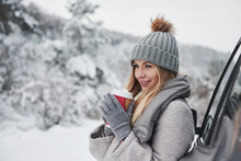 Warming Up By Fresh Beverage. Pretty Girl In Warm Clothes Standing In The Winter Wood While Leans On The Car And Holding Cup Of Coffee