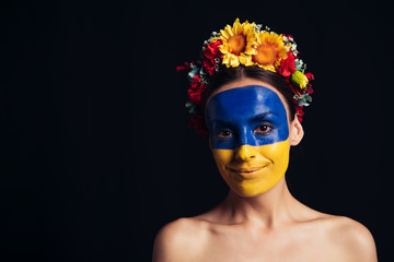 Wall Mural - naked young woman in floral wreath with painted Ukrainian flag on skin smiling isolated on black