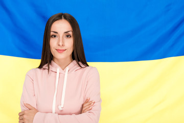 Wall Mural - smiling brunette young woman with crossed arms standing on Ukrainian flag background