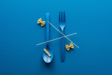 Wall Mural - top view of spaghetti on blue plastic spoon upside down and fork near two kinds of pasta on blue background