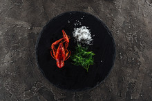 Top View Of Black Plate With Red Lobsters, White Salt And Green Herbs On Grey Textured Surface