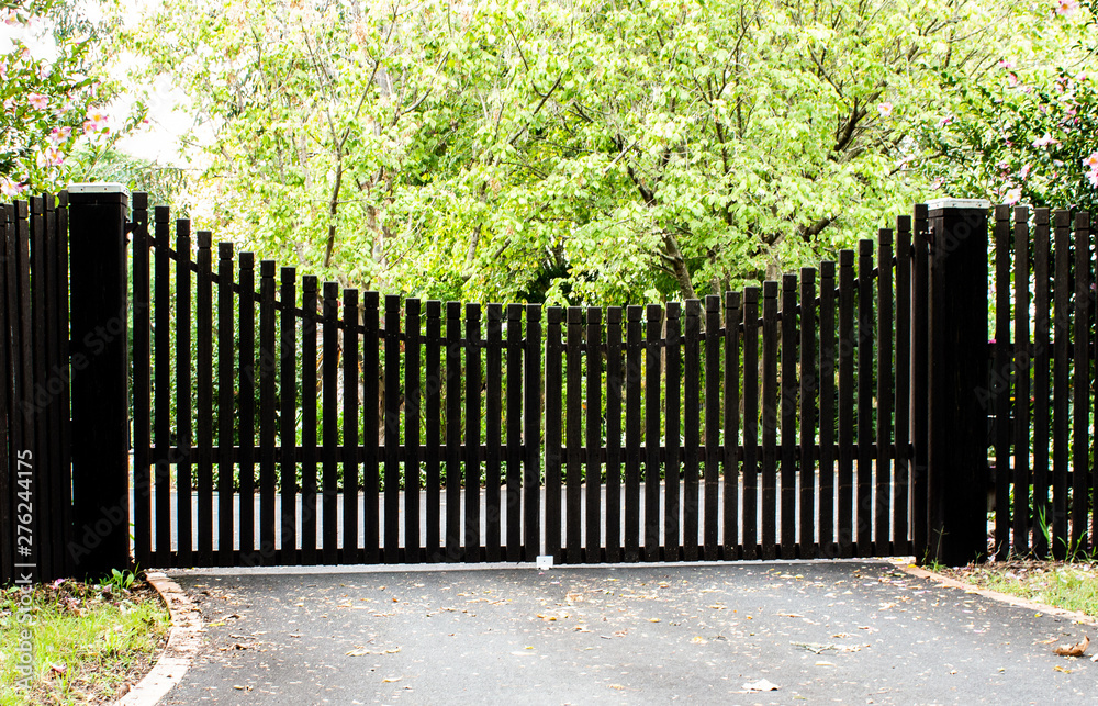 Dark wooden driveway property entrance gates set in timber picket fence with garden shrubs and trees in background - obrazy, fototapety, plakaty 