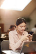© Seventyfour - Warm-toned portrait of young Asian student using smartphone while working at table in cafe, copy space