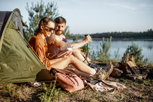 Young And Cheerful Couple Having A Picnic At The Campsite While Traveling In The Forest Near The Lake During The Sunset
