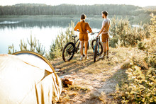 Young Couple Standing With Mountain Bicycles At The Campsite, Traveling In The Forest Near The Lake On The Sunset