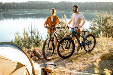 Young couple standing with mountain bicycles at the campsite, traveling in the forest near the lake on the sunset