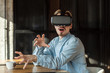 © khosrork - Portrait of suprised bearded young adult man in denim blue shirt sitting in cafe, wearing vr and watching video on simulator with raised arms and opened mouth. Indoor, lifestyle