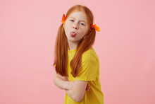 Portrait Of Petite Freckles Red-haired Girl With Two Tails, Looks And Shows Tounge At The Camera, Wears In Yellow T-shirt, Stands Over Pink Background.