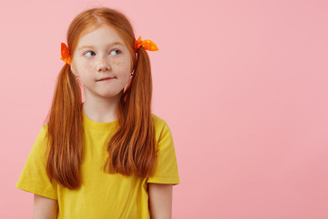 Wall Mural - Photo of little thinking freckles red-haired girl with two tails, looks away, touches cheeks, wears in yellow t-shirt, stands over pink background with copy space.