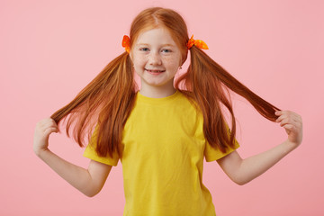 Wall Mural - Happy petite freckles red-haired girl takes his two tails, broadly smiling and looks cute, wears in yellow t-shirt, stands over pink background.