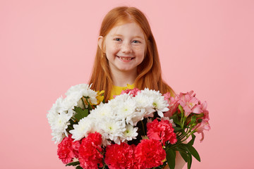 Wall Mural - Happy petite freckles red-haired girl with two tails, broadly smiling and looks cute, wears in yellow t-shirt, holds bouquet and stands over pink background.