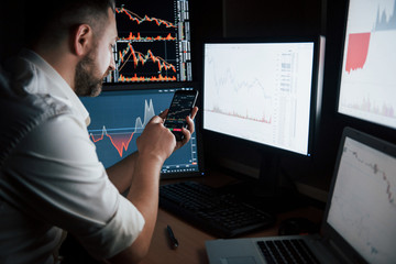 Wall Mural - New modern digital technologies. Bearded man in white shirt works by using smartphone in the office with multiple computer screens in index charts