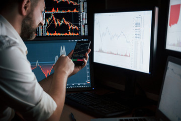 Wall Mural - Satisfied by results. Bearded man in white shirt works by using smartphone in the office with multiple computer screens in index charts