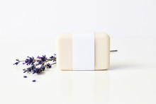Closeup Of Hand Made Herbal Soap Bar In Blank Paper Label Package And Bunch Of Lavender Flowers On White Table Backround. Spa Concept. Skin Product Mockup Scene. Cosmetic Product.