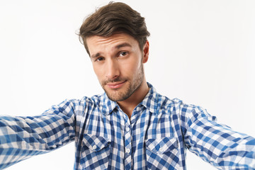 Canvas Print - Serious young unshaved man dressed in casual shirt posing isolated over white wall background take a selfie by camera.