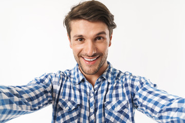 Canvas Print - Happy young unshaved man dressed in casual shirt posing isolated over white wall background take a selfie by camera.