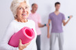 © Photographee.eu - Happy senior woman holding pink joga mat, men exercising in background