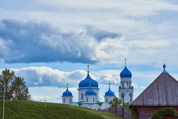 Beautiful white Church with blue domes against the cloudy sky. In the foreground is a green hill and a village house.