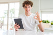 © Krakenimages.com - Young man using touchpad tablet doing ok sign with fingers, excellent symbol