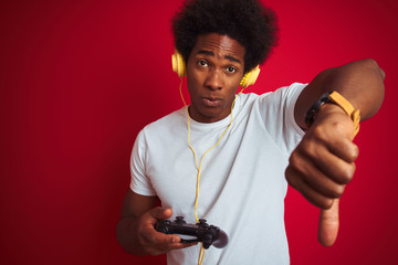 Afro gamer man playing video game using joystick headphones over isolated red background with angry face, negative sign showing dislike with thumbs down, rejection concept