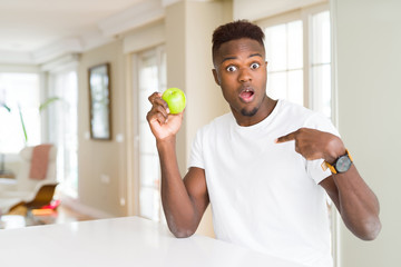 Young african american man eating fresh green apple with surprise face pointing finger to himself