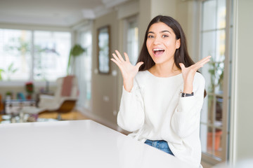 Young beautiful woman at home on white table celebrating crazy and amazed for success with arms raised and open eyes screaming excited. Winner concept