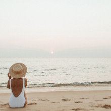 Summer Vacation Fashion Concept. Young, Tanned Woman Wearing A Beautiful White Swimsuit With A Straw Hat Is Sitting And Relaxing On Tropical Beach With White Sand And Is Watching Sunset And Sea.