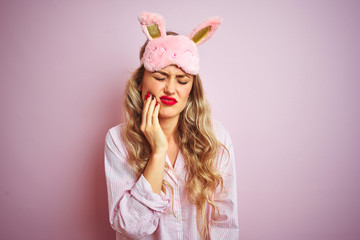 Young beautiful woman wearing pajama and sleep mask over pink isolated background touching mouth with hand with painful expression because of toothache or dental illness on teeth. Dentist concept.