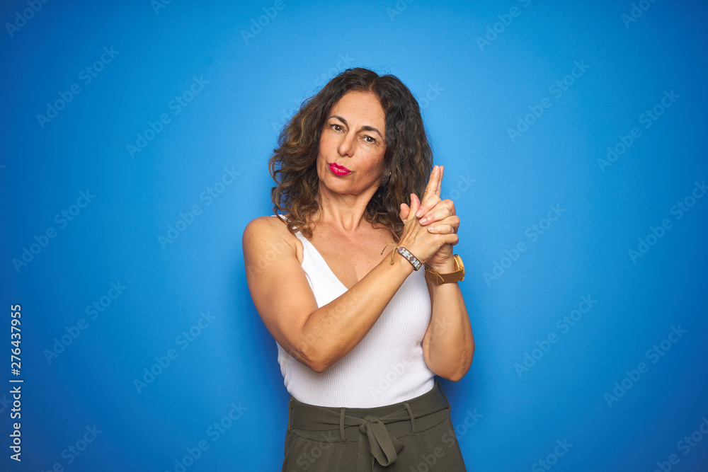 Middle age senior woman with curly hair standing over blue isolated background Holding symbolic gun with hand gesture, playing killing shooting weapons, angry face