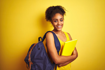 American student woman wearing backpack holding notebook over isolated yellow background with a happy face standing and smiling with a confident smile showing teeth