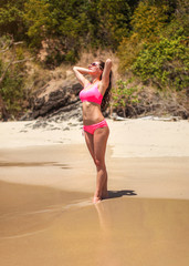 Wall Mural - Young sporty woman in pink bikini swimwear and sunglasses posing on the wet sand beach looking to sun, hands in her hair, jungle bushes behind