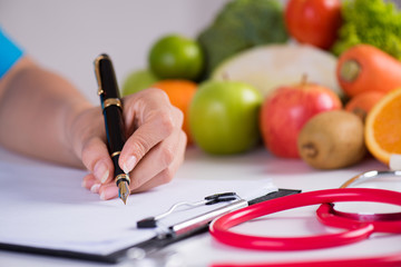 Healthy lifestyle, food and nutrition concept. Close up doctor woman hand holding pen to checklist with fresh vegetables and fruits with stethoscope lying on desk in background.