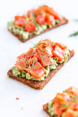 Wall Mural - Close-up view of three sandwiches with rye bread, avocado and smoked salmon on a white kitchen table.