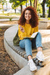 Young beautiful curly student girl sitting outdoors in nature park writing notes in notebook.