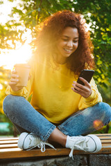 Wall Mural - Happy cheerful smiling optimistic cute young student curly girl sitting on bench outdoors in nature park with beautiful sunlight using mobile phone.