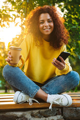 Wall Mural - Happy optimistic cute young student curly girl sitting on bench outdoors in nature park with beautiful sunlight using mobile phone.