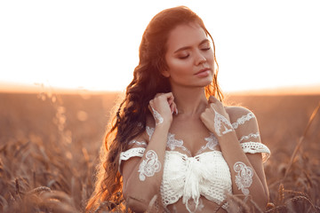 Boho chic style. Portrait of bohemian girl with white art posing over wheat field enjoying at sunset. Outdoors photo. Tranquility concept. Lifestyle.