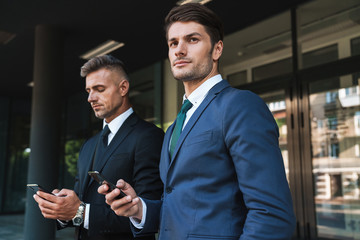 Wall Mural - image of caucasian businessmen partners standing outside office center and holding cellphones togeth