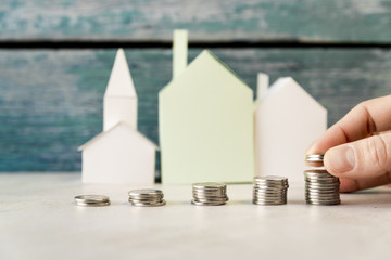 A person arranging the increasing coins in front of paper houses on white surface