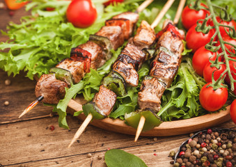 Grilled pork and chicken kebab with paprika in round wooden plate of lettuce salad, on wooden background with tomatoes and spinach. Macro