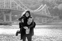 Beautiful Amazing Funny Cheerful Young Couple (man And Woman) Outdoor By The River On Bridge Background. Girlfriend Riding On Boyfriend Back. Family, Love And Friendship Concept