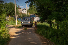 A Minibus Moving The Dirt Road Near House, Trees. A Girl Walkin From The Beach.
