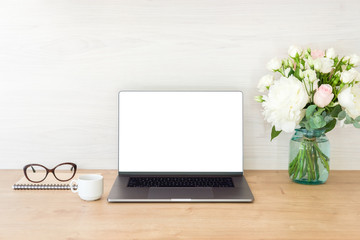 Office desk table with laptop computer with blank screen mockup and flowers bouquet, coffee cup, glasses, notepad on wooden surface. Minimal style feminine workplace concept.