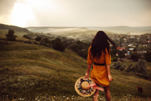 Back View Of A Young Woman With Dark Long Hair Holding A Hat And Walking Down The Hill Against Sunset.