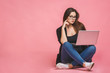 Business concept. Portrait of happy woman in casual sitting on floor in lotus pose and holding laptop isolated over pink background