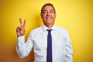 Handsome middle age businessman standing over isolated yellow background showing and pointing up with fingers number two while smiling confident and happy.