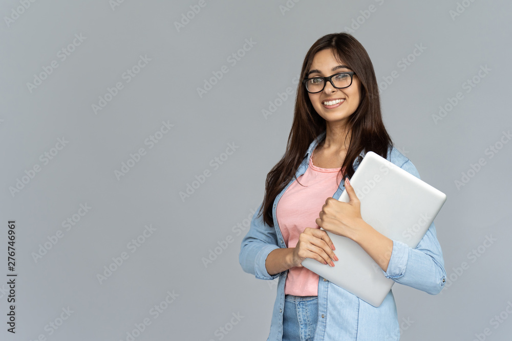 Smiling indian young woman college university school student wear glasses holding laptop computer isolated on grey studio background, happy teen girl looking at camera stand with device, portrait