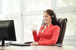 © spaskov - Young attractive woman in red jacket sitting on desk at head of executive in red jacket. Holds video conference in headphones with microphone. Drinking coffee from white cup.