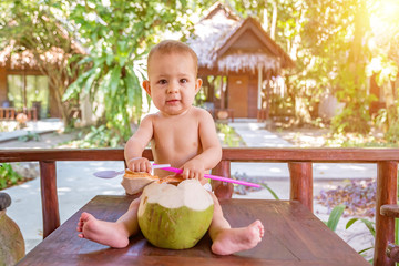 teething and salivation infant baby at tropical vacation. Eats and drinks green young coconut. Sits on a table